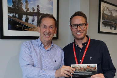 Colin Earps and Andy Pendlebury smiling while holding a certificate or printed image, standing in front of framed landscape photographs on the wall.